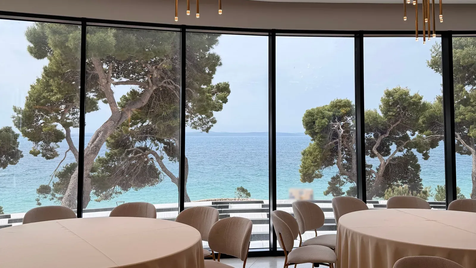 Round tables with cream cloths and upholstered chairs fill an event room whose floor-to-ceiling windows look out on pine trees and the sea.