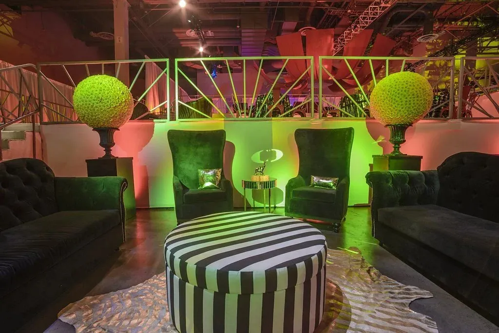 Lounge seating with green velvet chairs and a striped ottoman under colored lighting