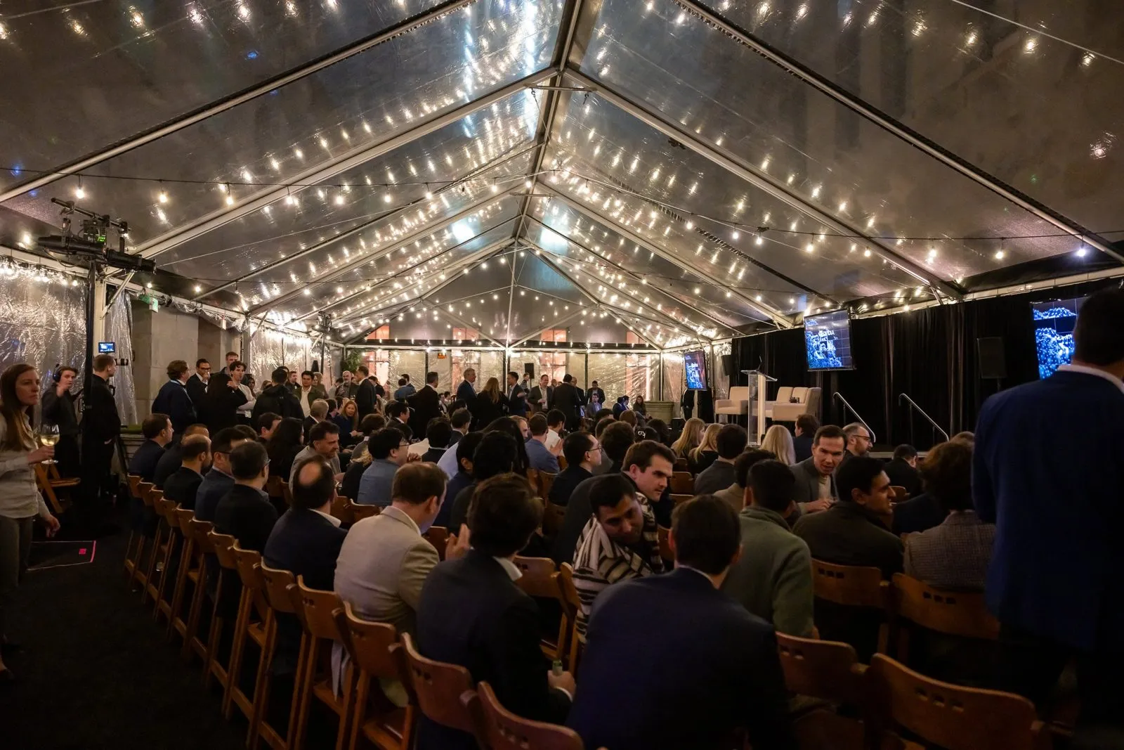 Seated audience under a clear-roof event tent with string lights
