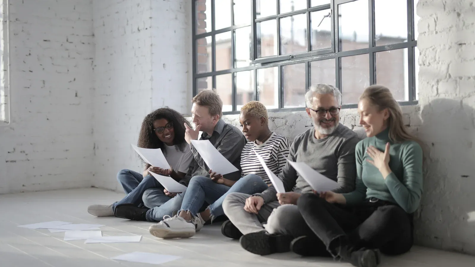Five colleagues sit on a sunlit floor against a white brick wall, reading printed pages beside a large window.
