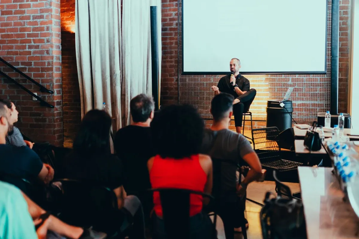 An audience seated in a brick-walled event space listens to a speaker with a microphone in front of a blank projection screen.