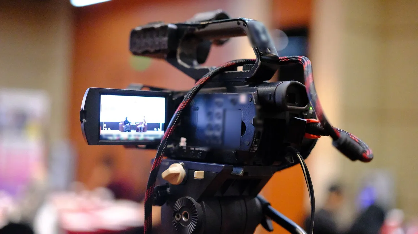 A video camera on a tripod records a panel on stage while the audience sits softly blurred behind it for a virtual sales kickoff.