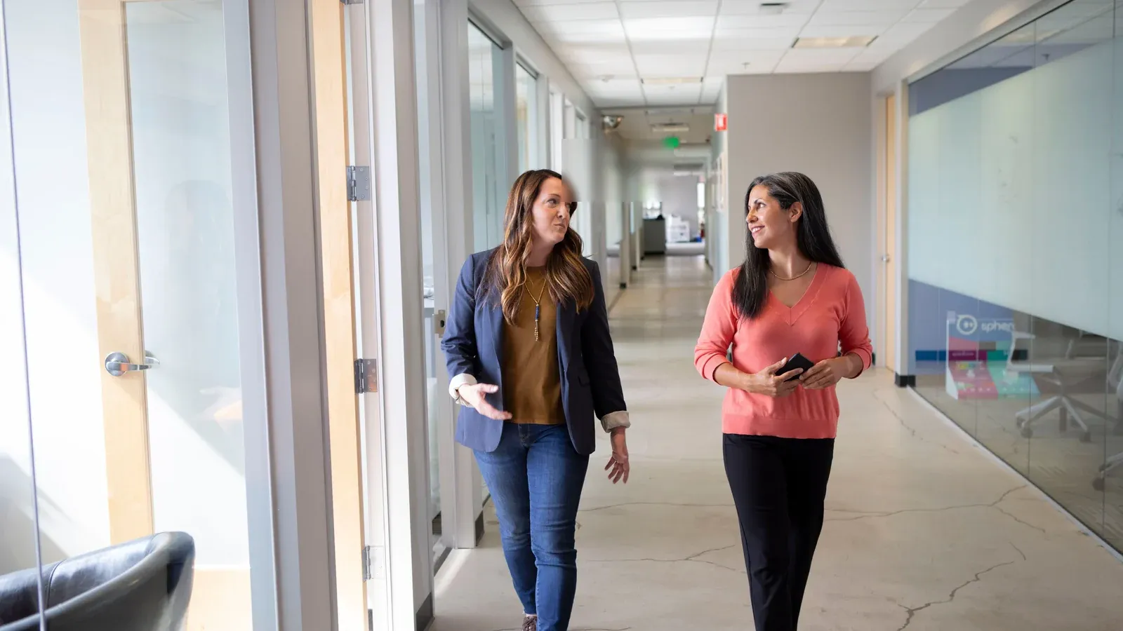 Two colleagues walk and talk down a bright office corridor. Glass walls and daylight line the hallway beside them.