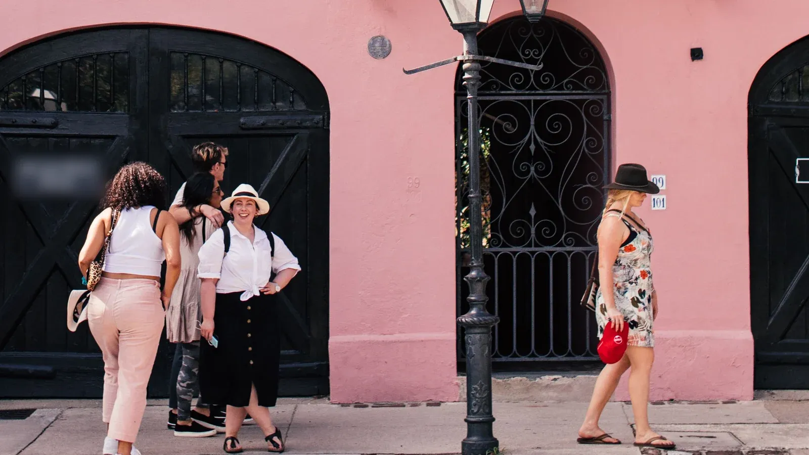 A small group pauses outside a pink historic building with black arched doors, an iron gate and a street lamp during an incentive trip in Charleston.