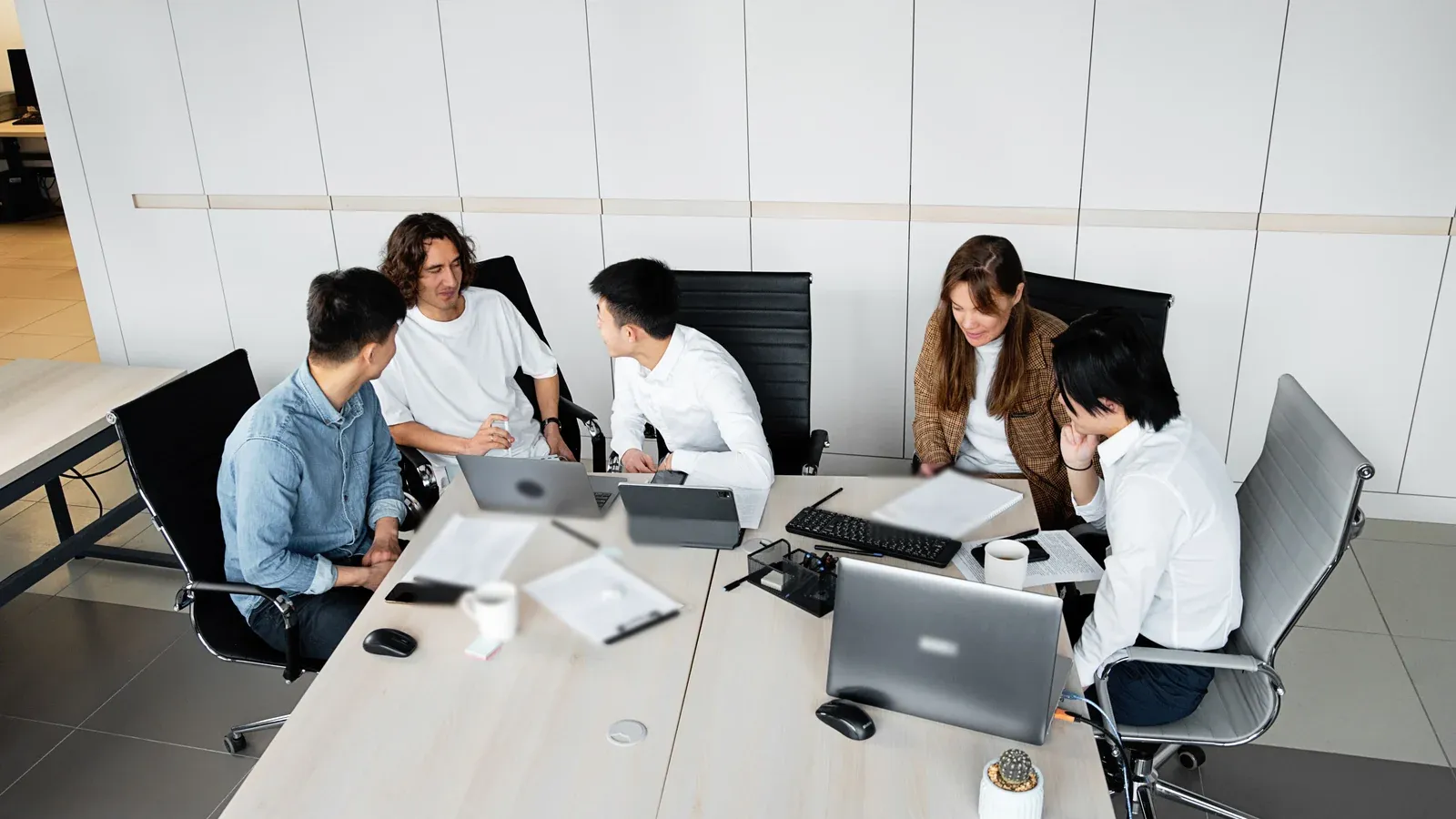 A high-angle view shows five colleagues gathered around joined white desks with laptops, notes and coffee cups in a bright office during a company offsite.