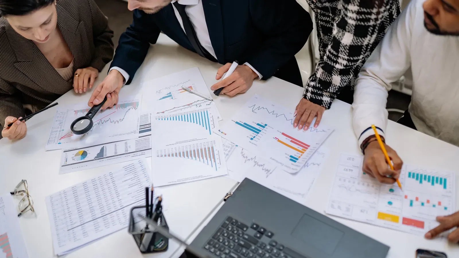 Overhead view of four colleagues marking up printed bar charts and spreadsheets beside a laptop on a white table.