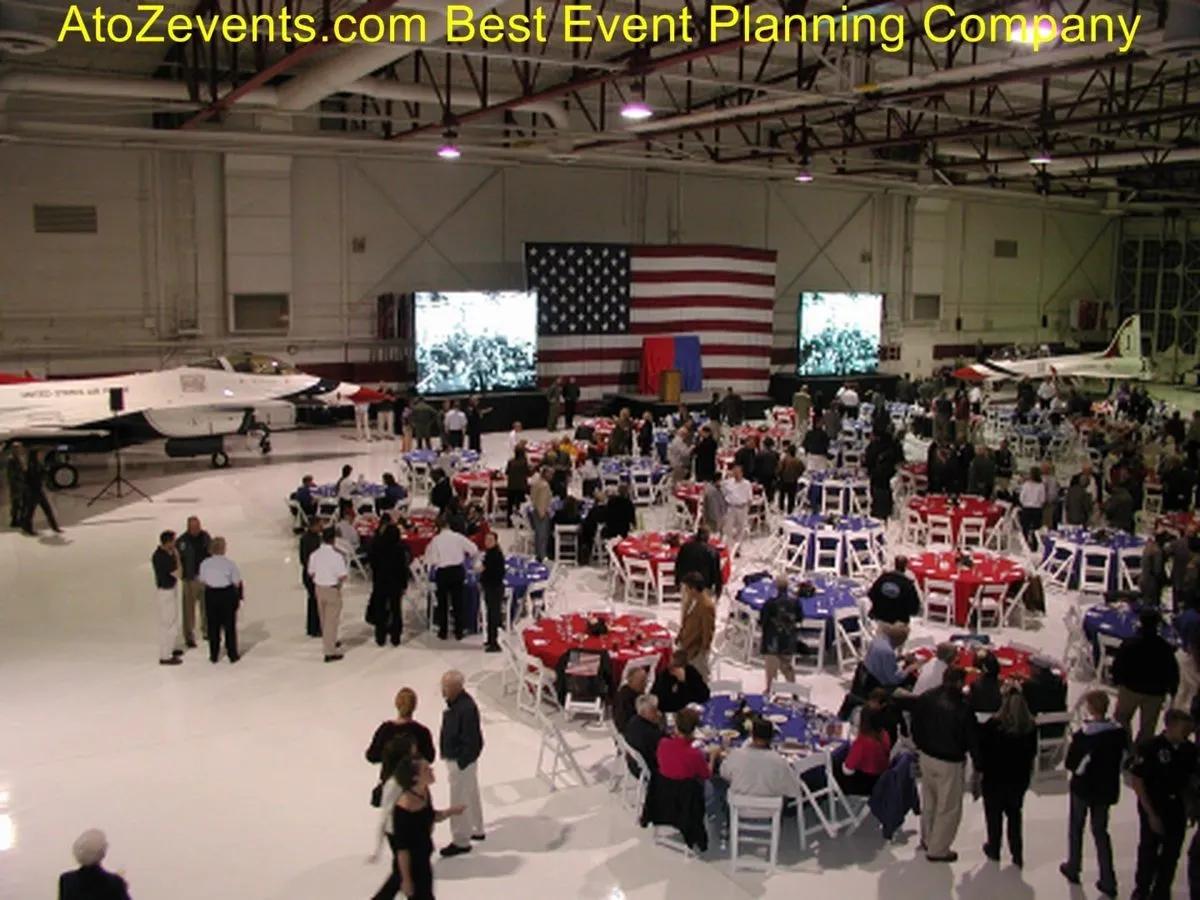 Guests at round tables in an aircraft hangar with screens and a US flag
