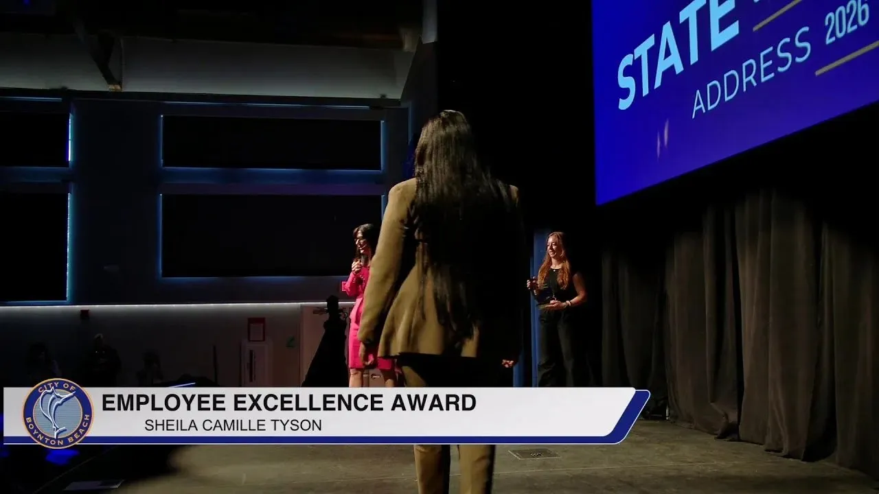 Award presentation on stage at the Boynton Beach State of the City, with broadcast lower-third graphics