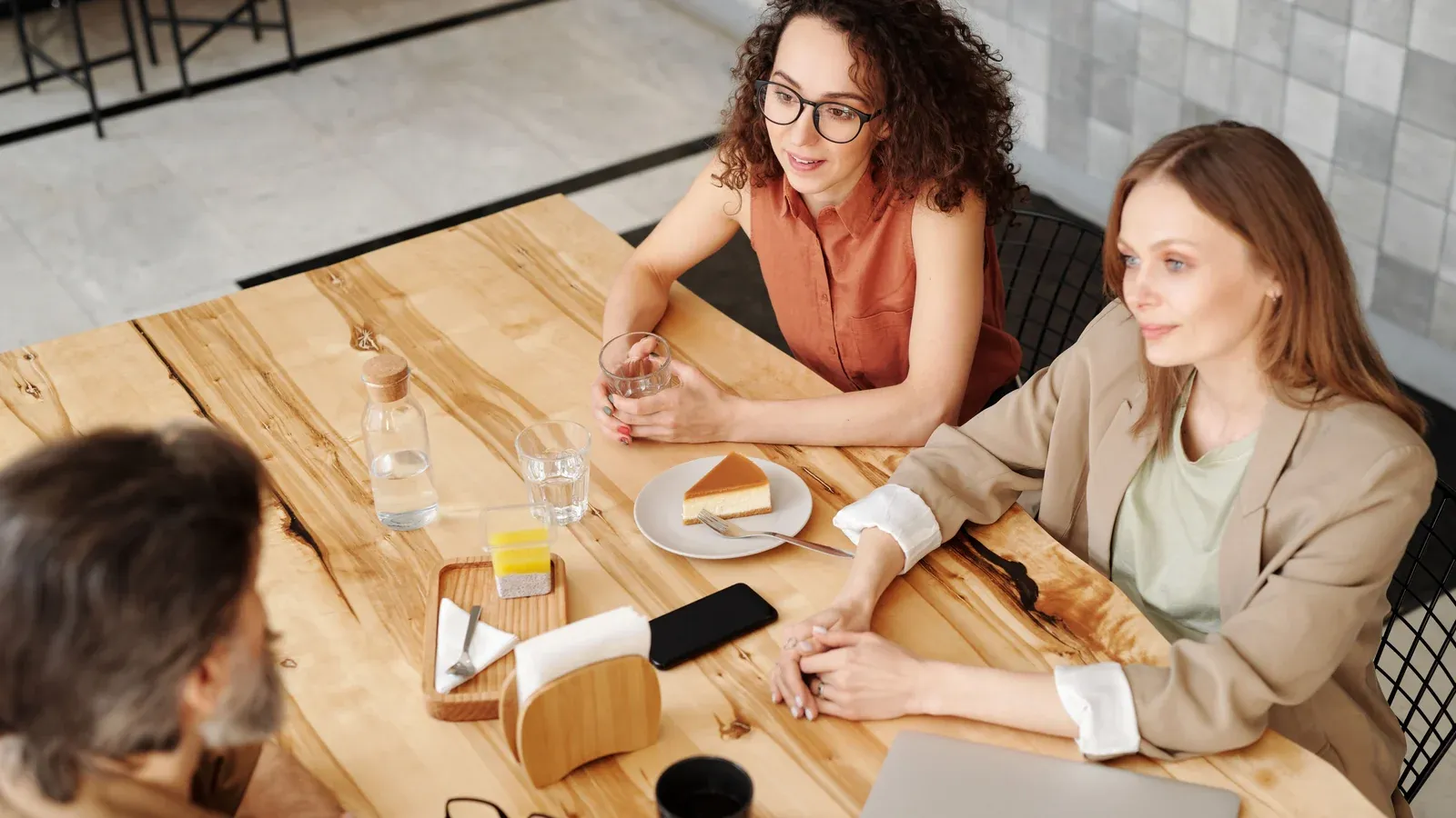 Three colleagues talk over coffee at a sunny wooden table. Two women listen closely as the conversation continues.