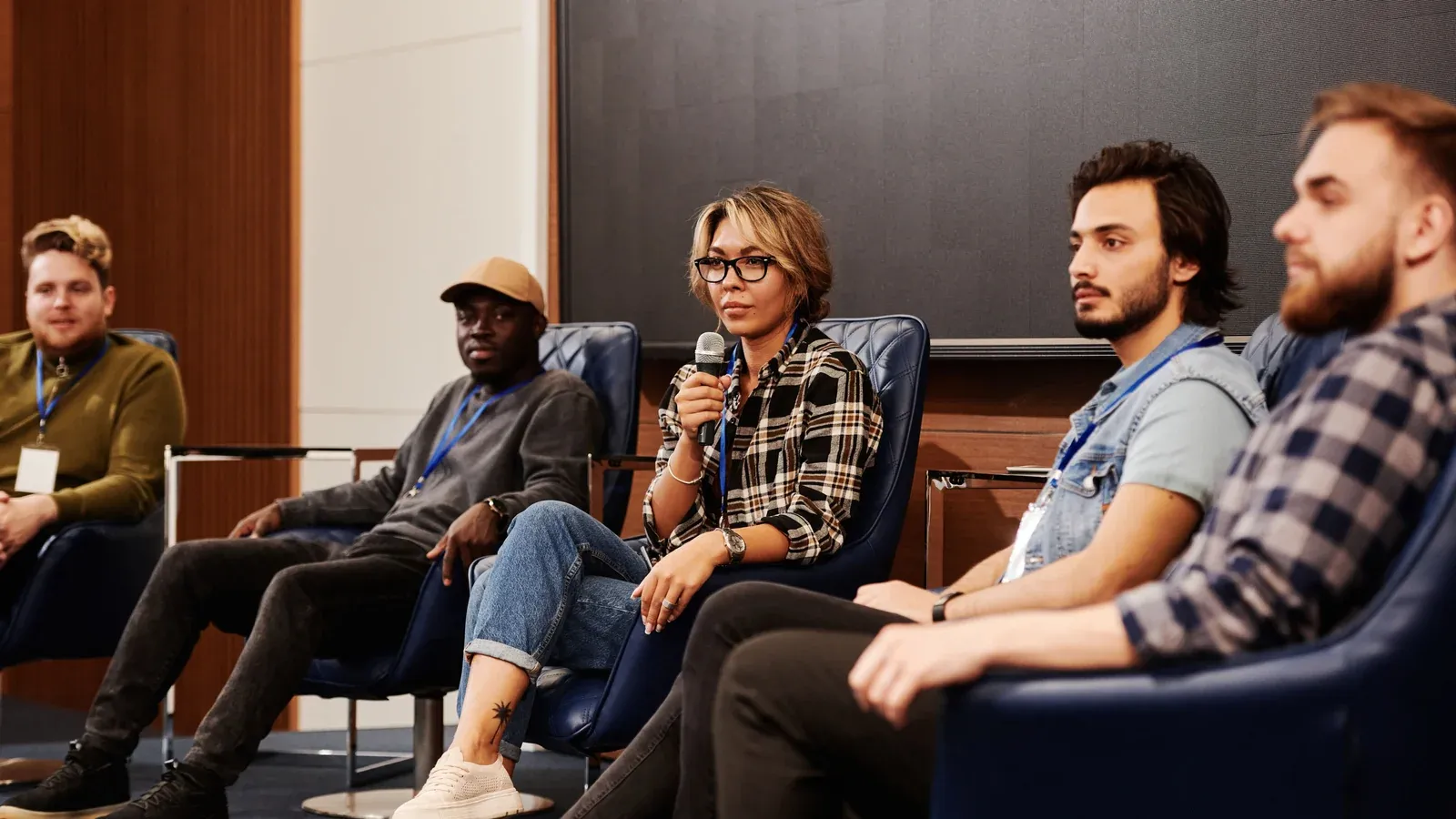 Five panelists sit side by side on low chairs during a conference discussion. One speaks into a microphone while the others listen.