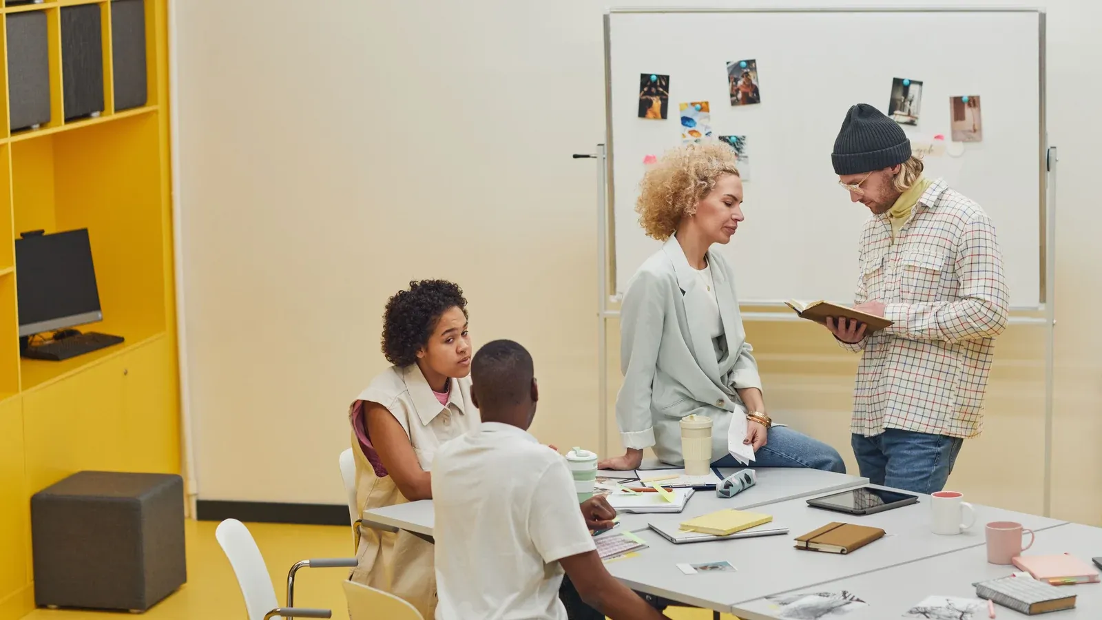 Two colleagues confer at a whiteboard pinned with photos while two teammates work at a shared table of notebooks and mugs in a bright room during a company offsite.