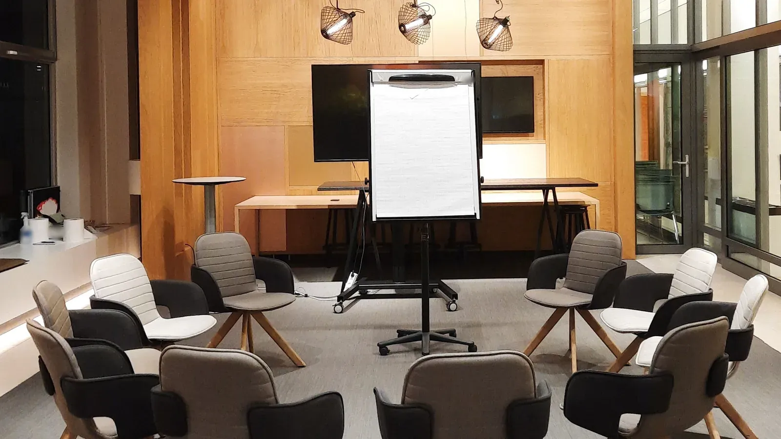Upholstered chairs are arranged in a loose circle around a blank flip chart in a warm, wood-panelled workshop room. The space is set and waiting for a team to arrive.