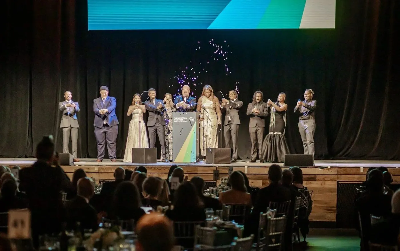 Group on stage applauding at an evening program with a large screen above