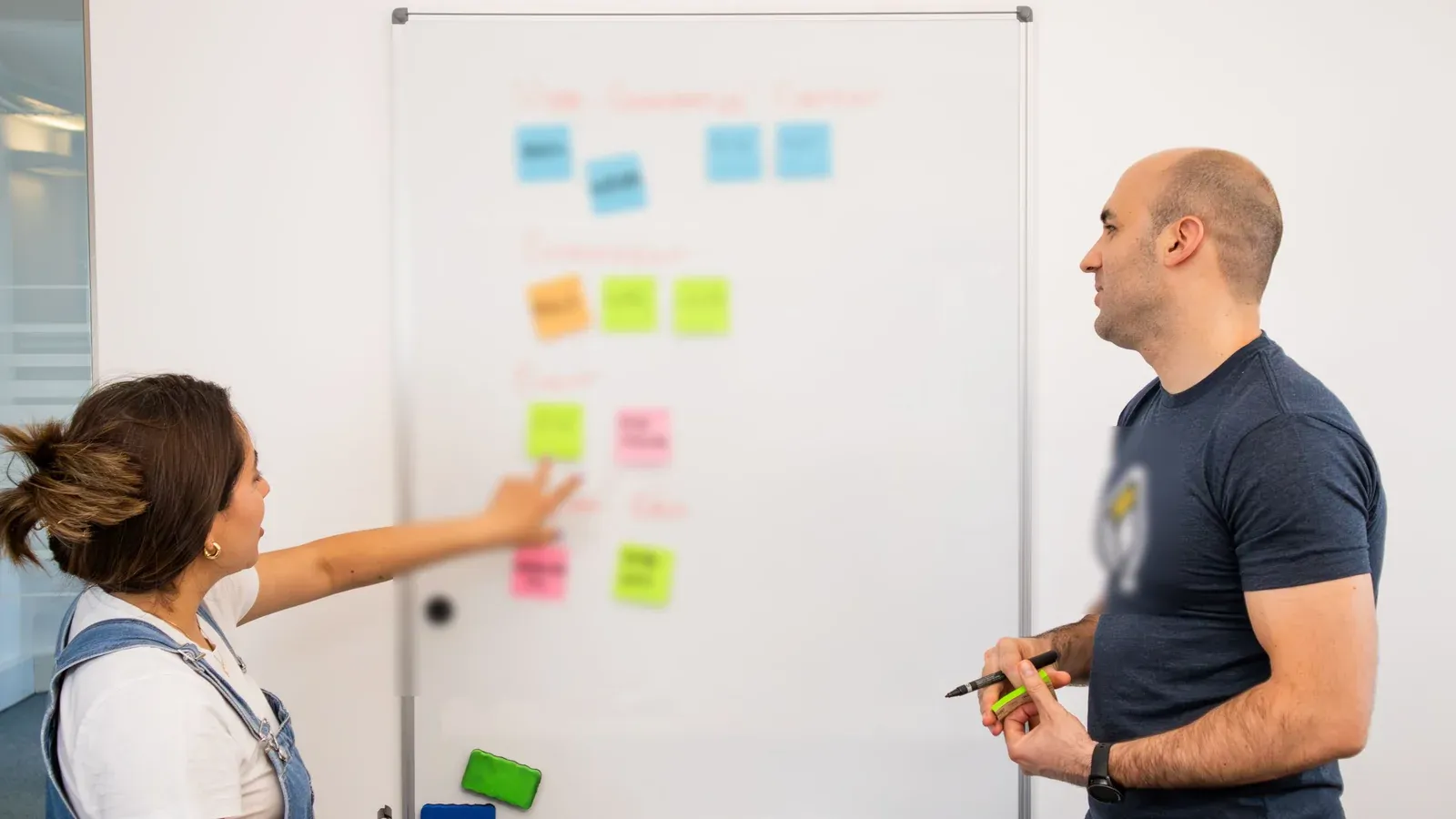 Two colleagues plan a project together at a whiteboard covered in sticky notes. One points to a note while the other listens with a marker in hand.
