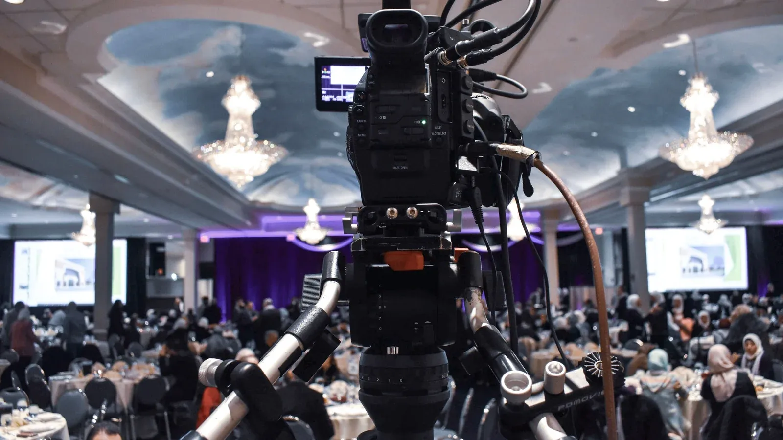 A video camera on a tripod films a large meeting in a hotel ballroom. Hundreds of attendees sit at round tables beneath crystal chandeliers.