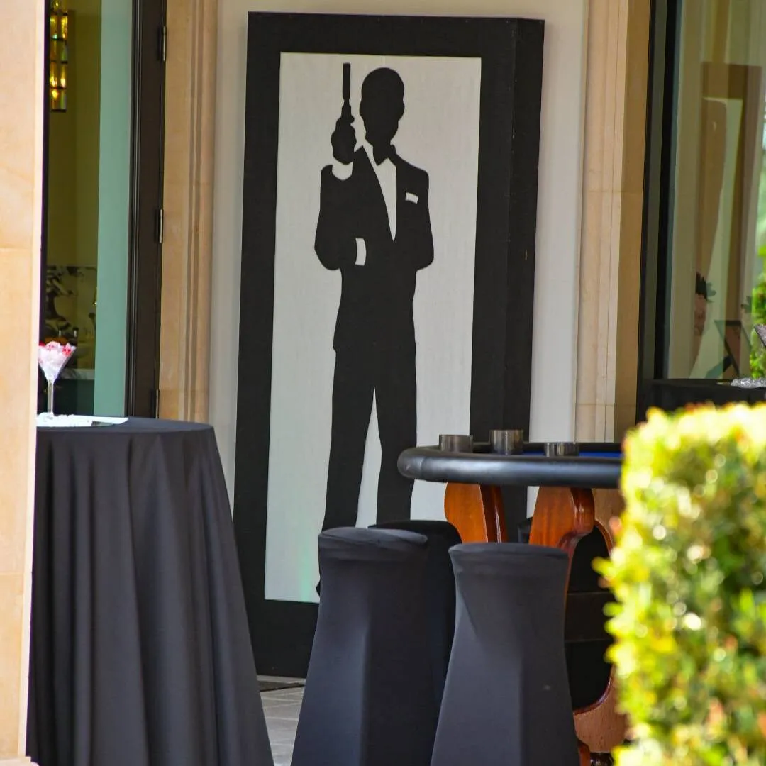 Casino-night decor with a tuxedoed silhouette panel, a card table and black-draped cocktail tables at the Impact 100 Men charity event, May 2024