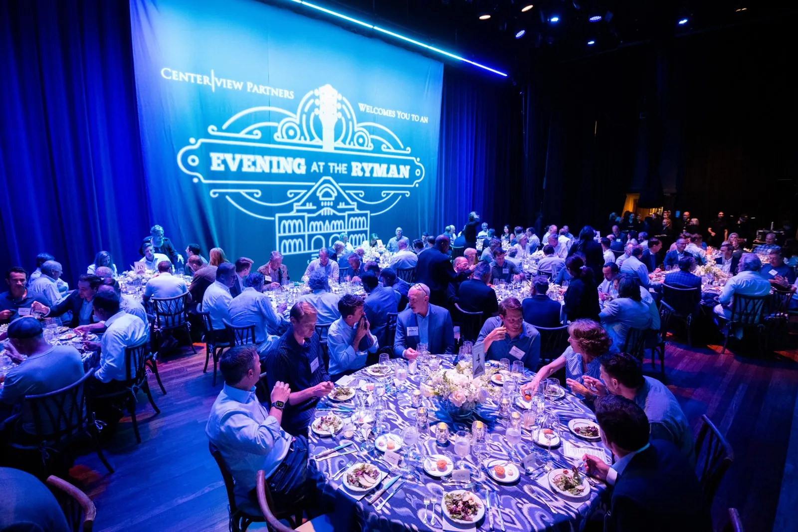 Guests at a seated dinner under blue lighting with a branded screen above the stage