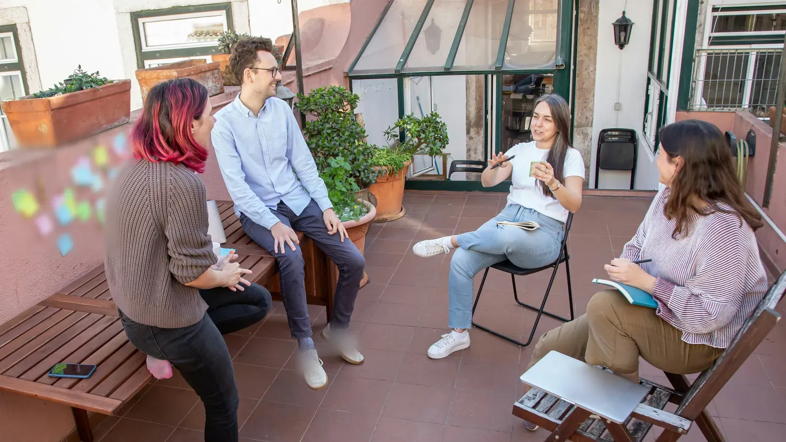 Four colleagues talk while seated on an outdoor terrace, one holding a notebook, with a closed laptop on a chair.