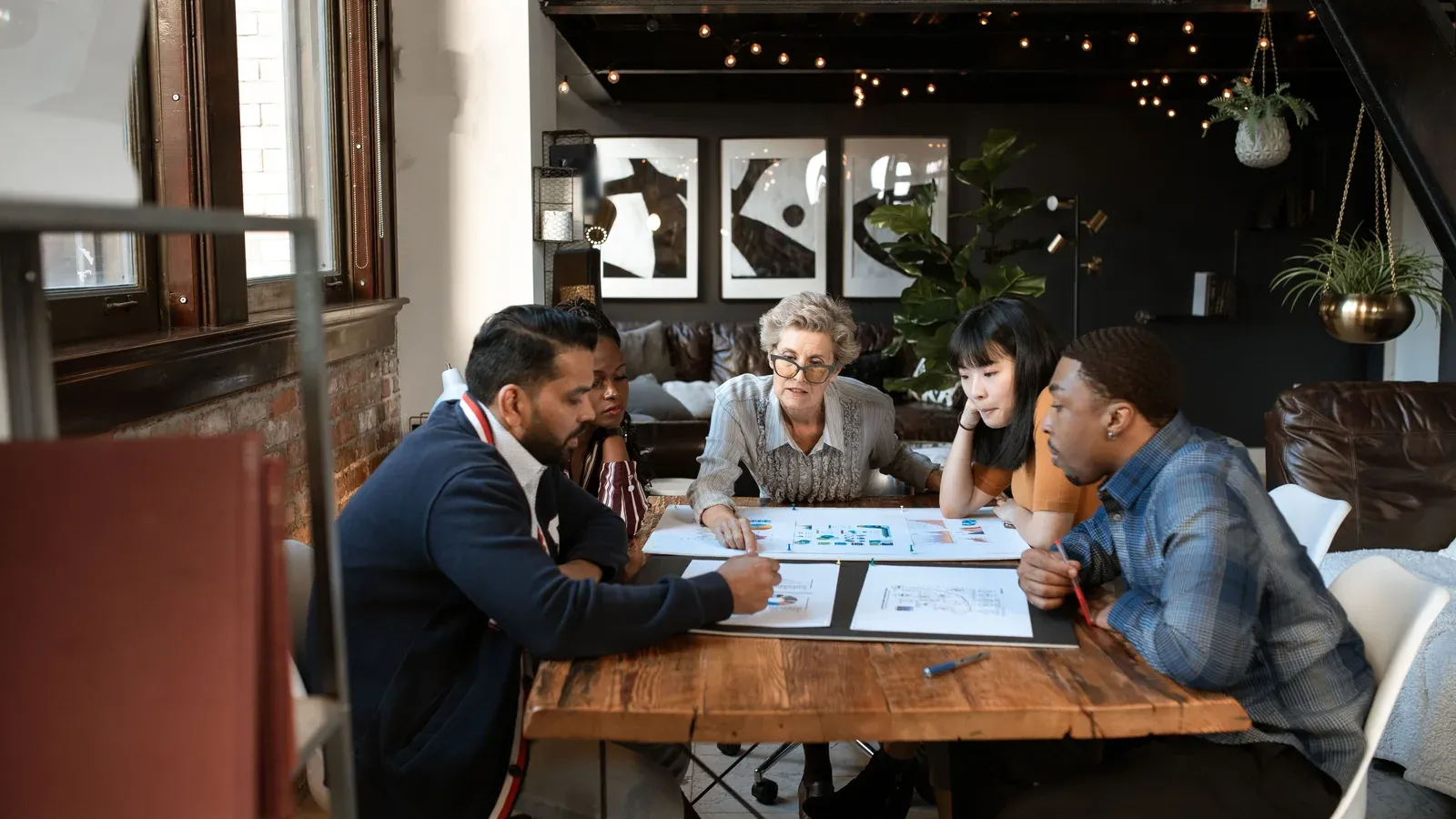 Five colleagues lean over printed charts on a wooden table in a loft lounge with brick walls and string lights.