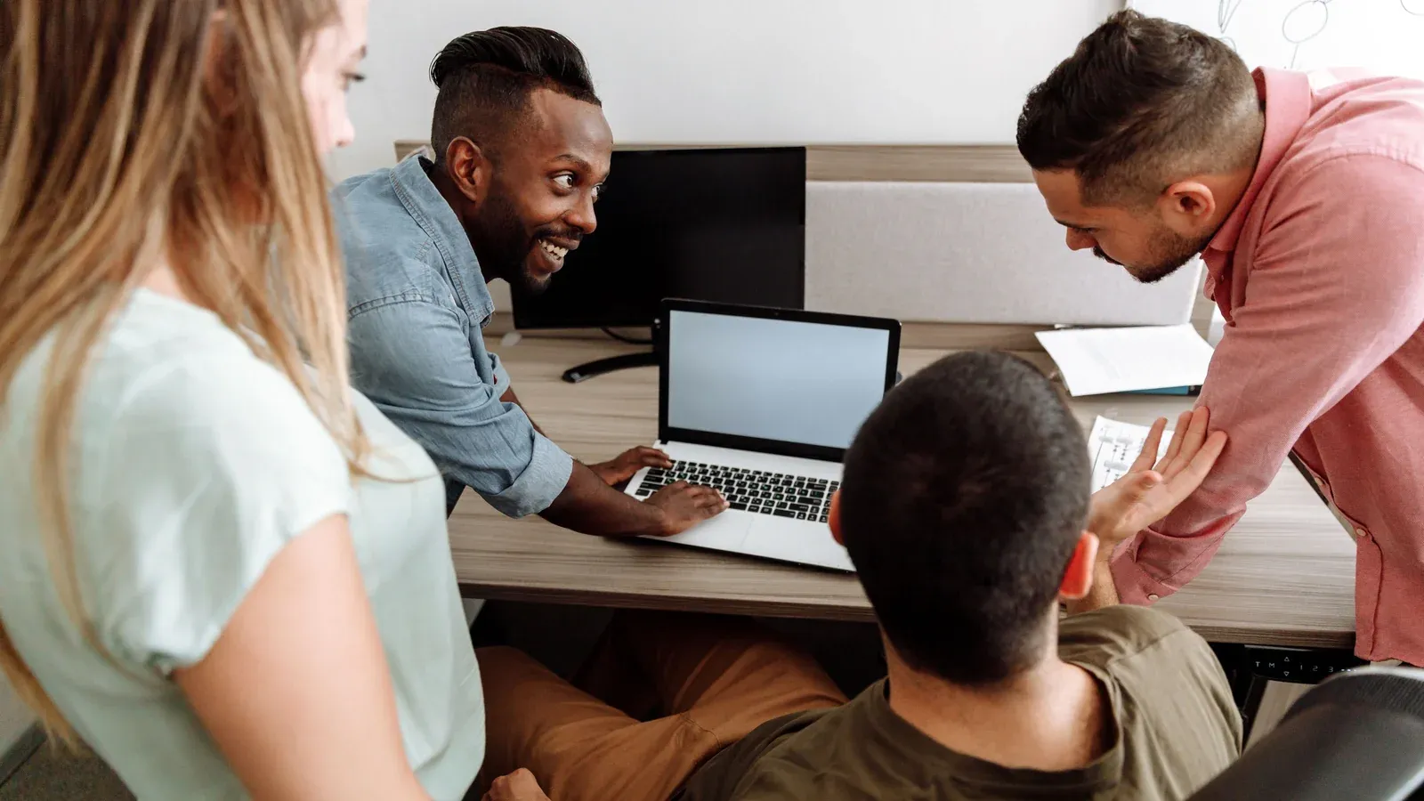 Four colleagues gather around a laptop at a shared desk in a bright office. One leans in to point at the screen while the others smile and follow along.