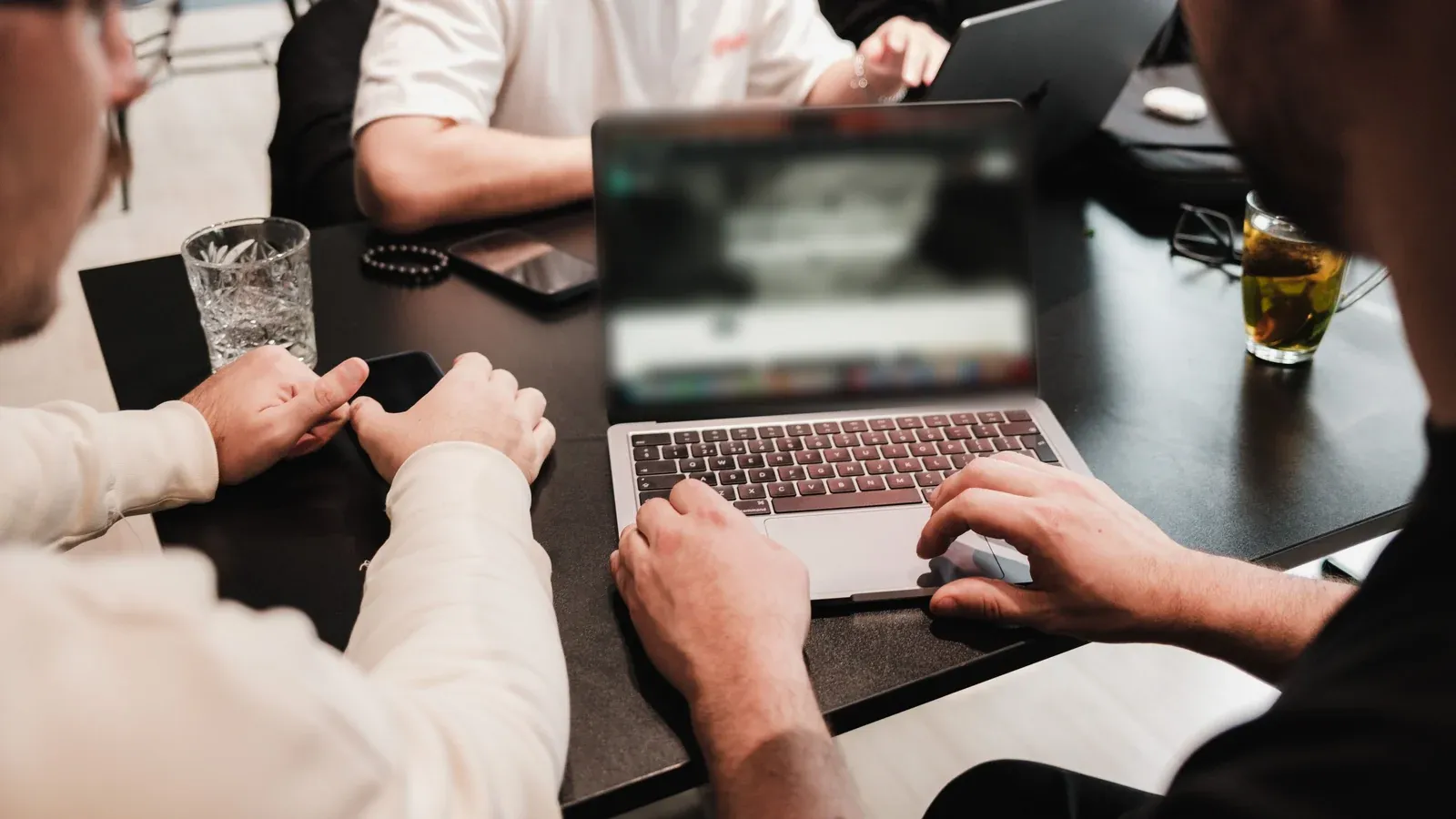 Teammates lean in over a shared laptop at a dark worktable with water and tea close at hand during a hands-on working session at a company offsite.