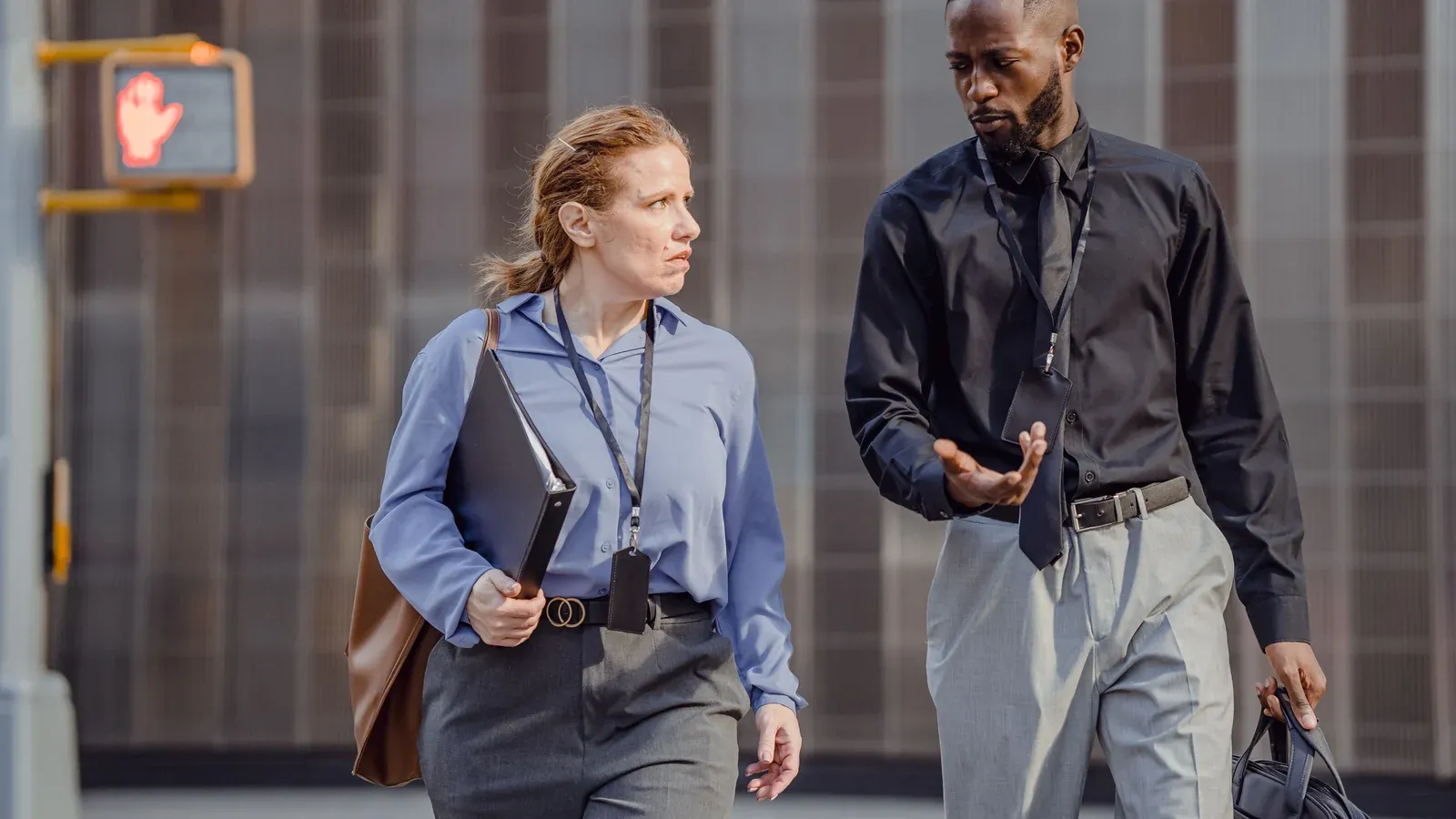 Two colleagues in business attire talk as they walk past a modern office building. Both carry work bags and wear conference lanyards.