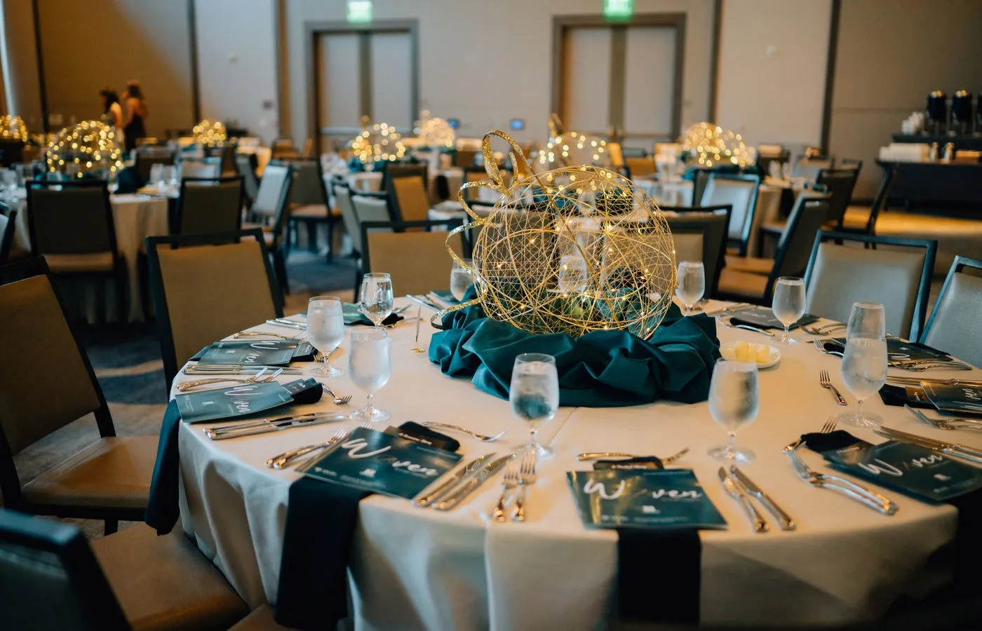 Round dinner tables with teal linens and gold centerpieces in a ballroom