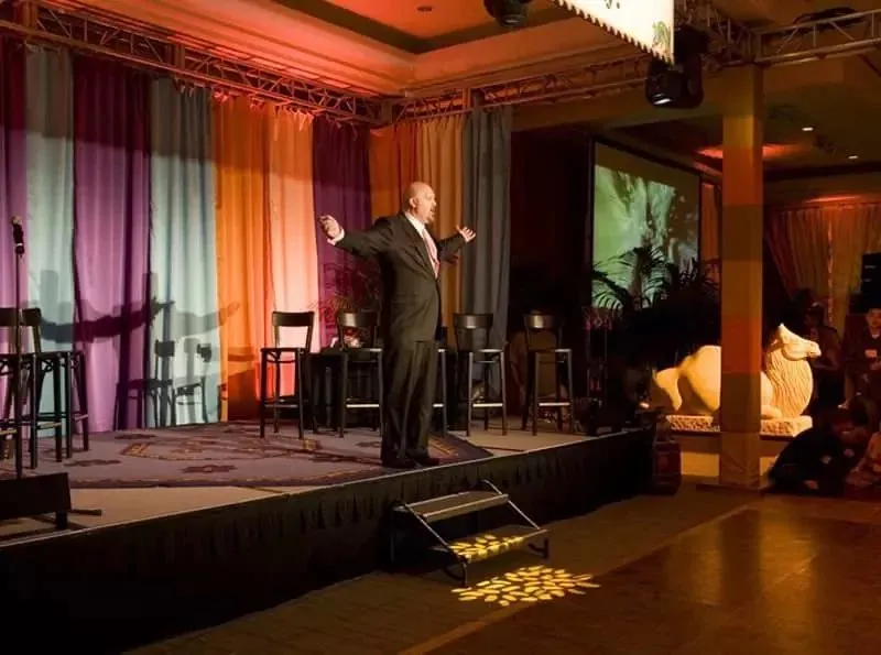 Speaker presenting on a small stage with draped backdrop and cocktail tables