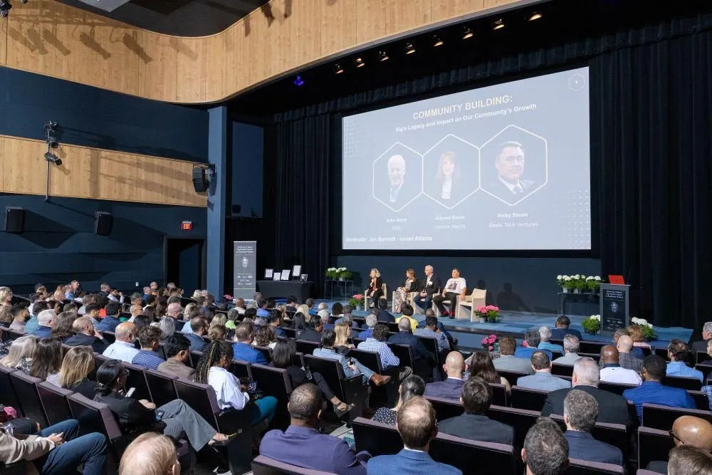 Conference audience seated in a theater facing a panel and presentation screen