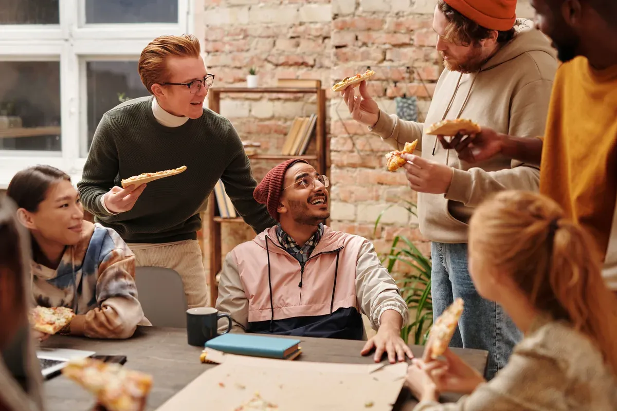 Colleagues crowd around a wooden table in a bright, brick-walled room, trading jokes over slices of pizza at a relaxed midday lunch. A daytime party can be as simple as stopping work to eat together.