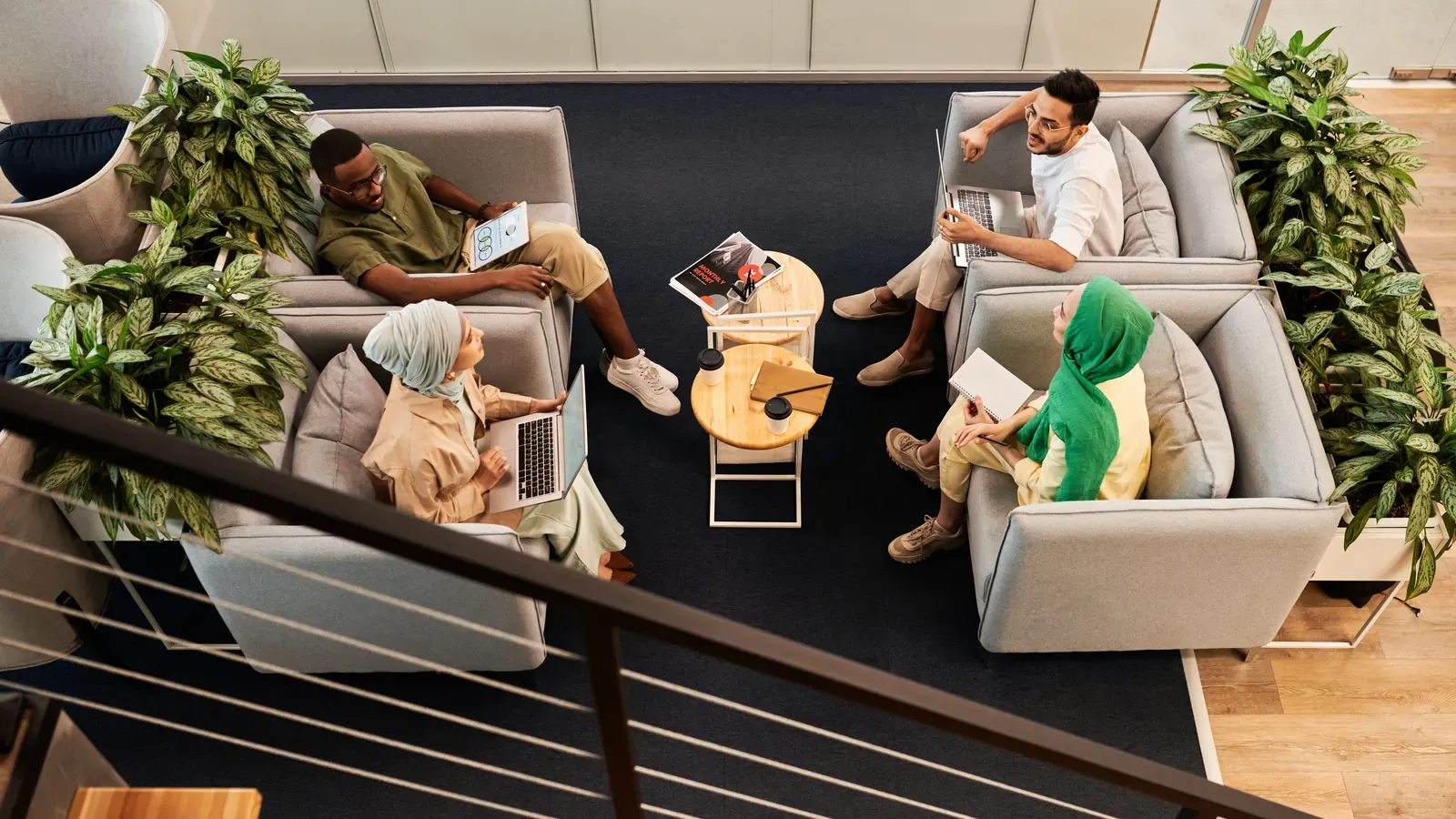 Seen from above, a team meets on sofas in a modern office lounge with laptops and notebooks. Green plants frame the relaxed discussion.