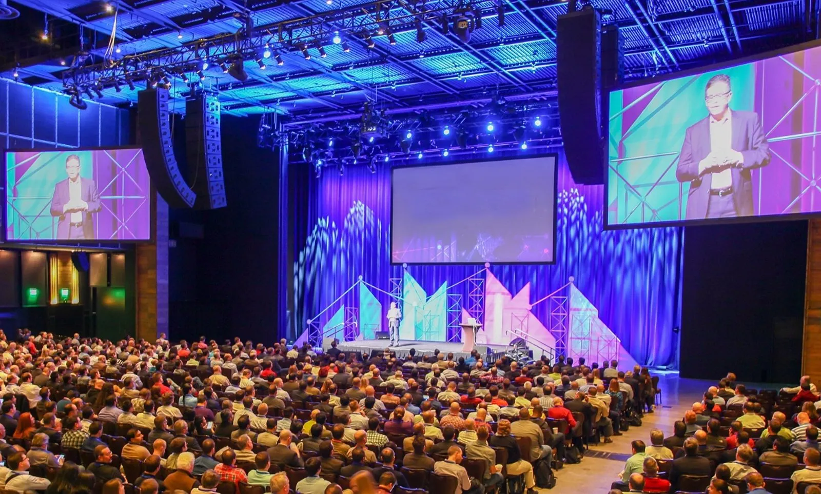 Large audience facing a lit conference stage with speaker and screens
