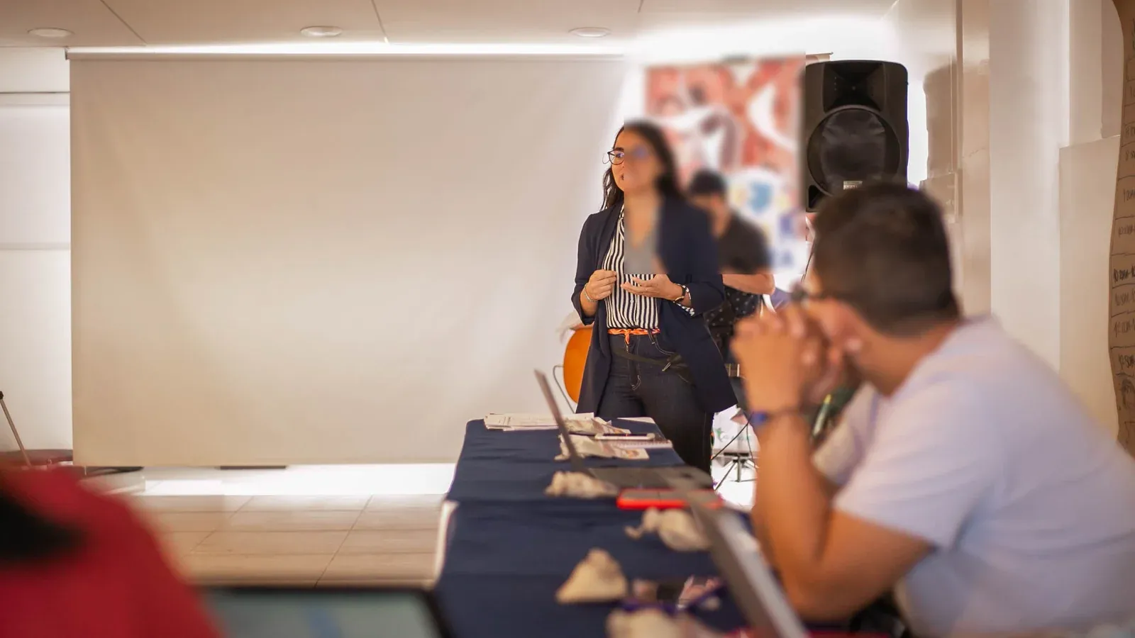 A woman stands at the front of a bright meeting room and speaks to her colleagues. Attendees seated along a long table listen and take notes.