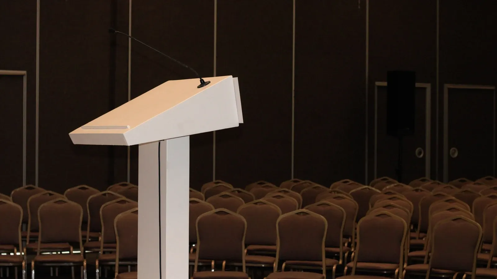 A white wooden lectern with a slim gooseneck microphone stands at the front of a dark conference hall. Rows of empty upholstered banquet chairs fill the room behind it, with a tall speaker on a stand near the back wall.