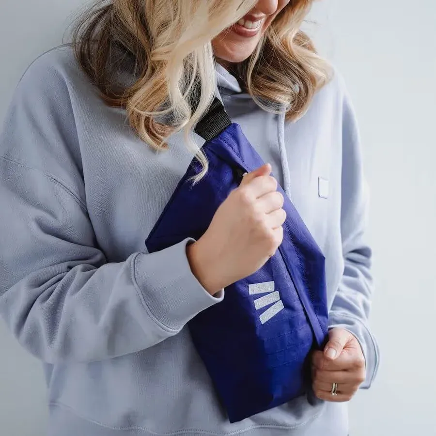 A woman in a light gray hoodie wears a royal blue nylon sling bag across her chest, with a small white printed logo near the bottom