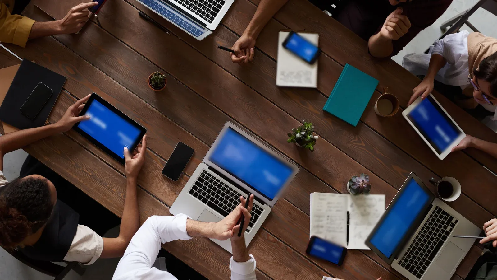 An overhead view shows a team gathered around a long wooden table with laptops, tablets and notebooks during a working session at a company offsite.