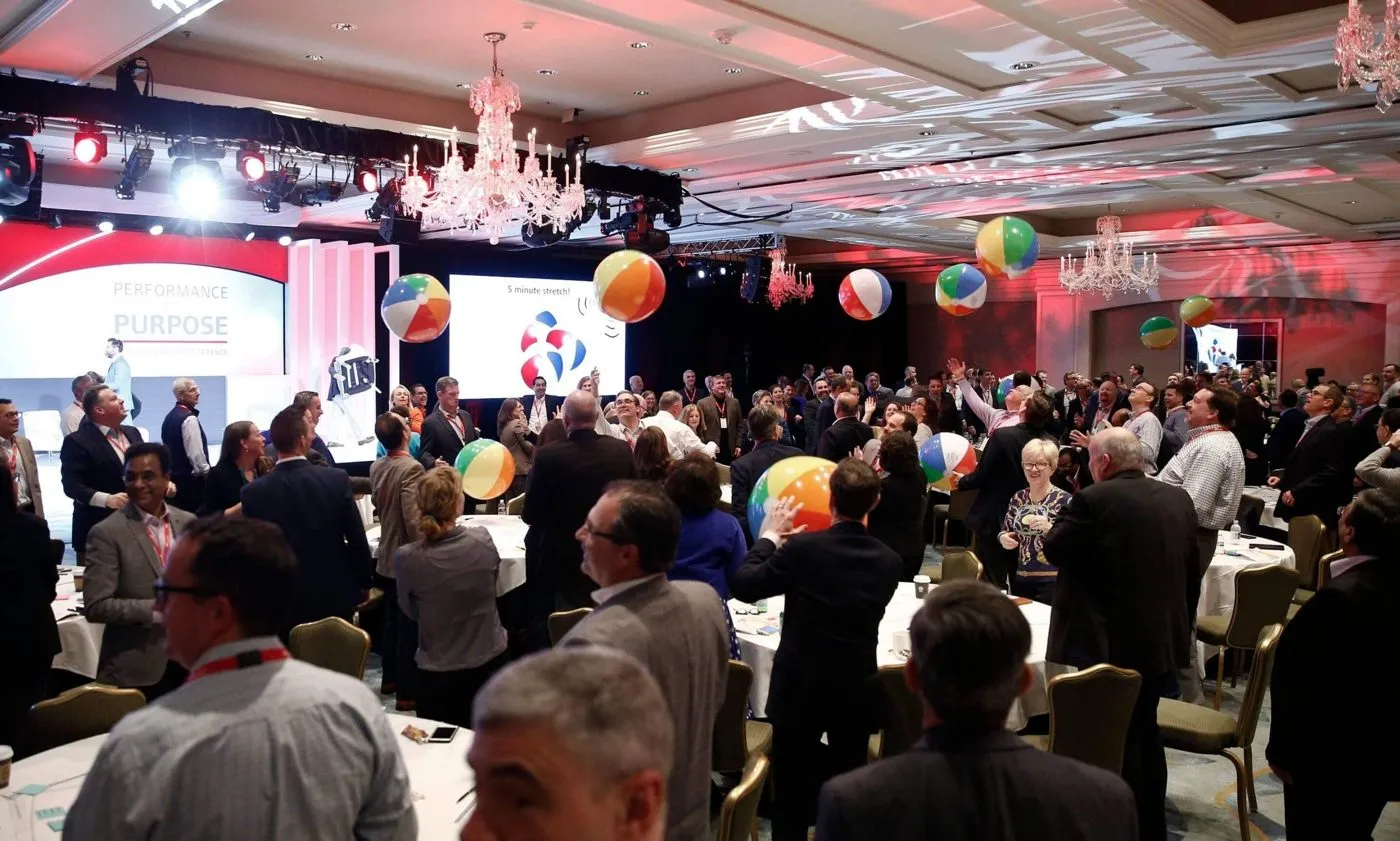 Large ballroom event with balloons, stage screens and a standing crowd