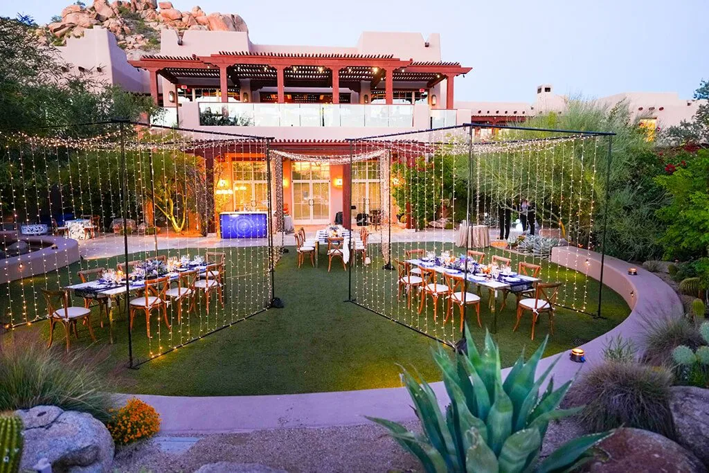 Dinner tables on a lawn framed by hanging lights beside a desert garden