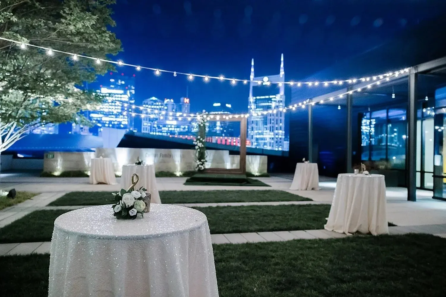 Cocktail tables and string lights on a lawn at dusk with a city view
