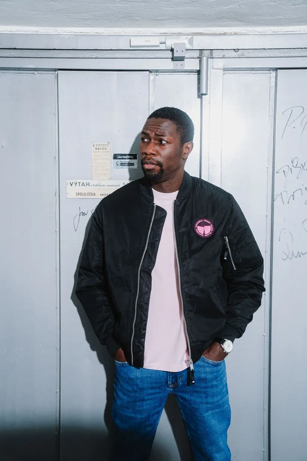A man in a black bomber jacket with a small round pink patch on the chest, worn open over a pale pink tee and jeans, stands against a gray metal door