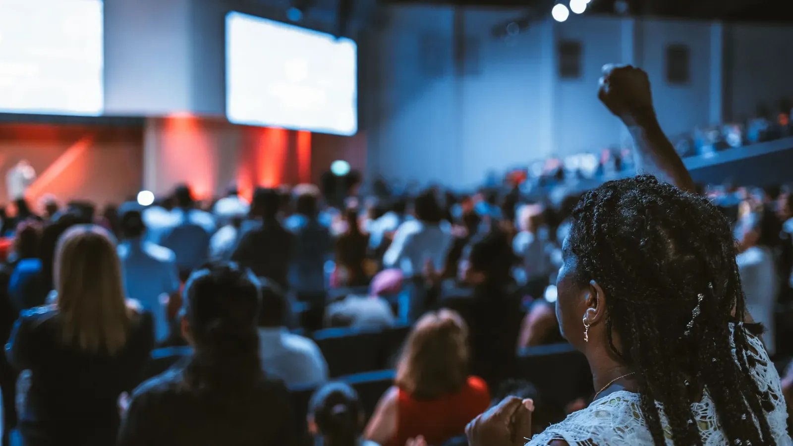 A large audience fills a dim event hall facing two bright stage screens while one attendee raises a fist.