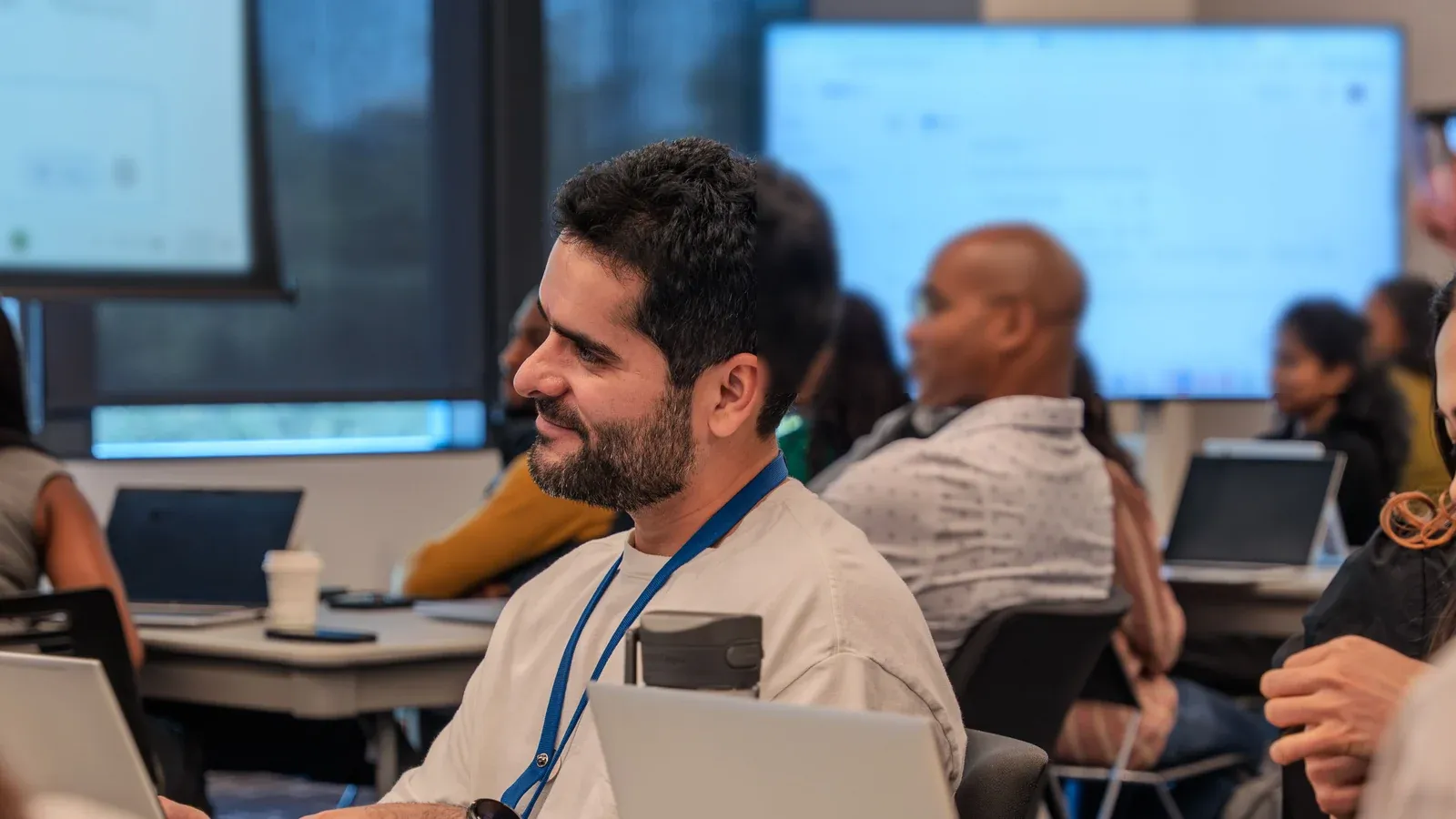 Participants with laptops sit at tables during a business training session. A man in a lanyard smiles as he listens to the speaker.