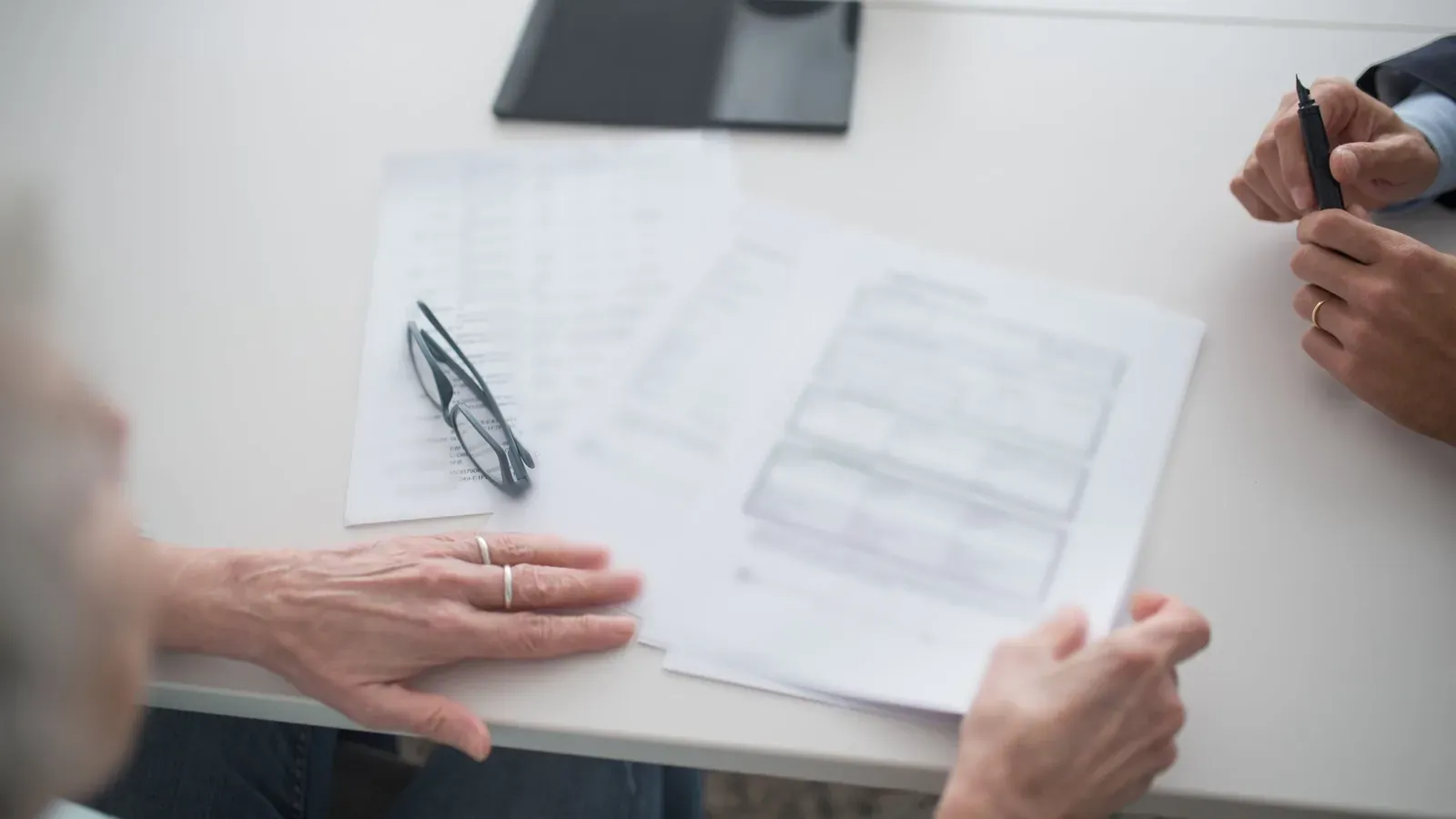 Hands rest beside printed forms, a pair of glasses and a pen on a pale meeting table during a document review.