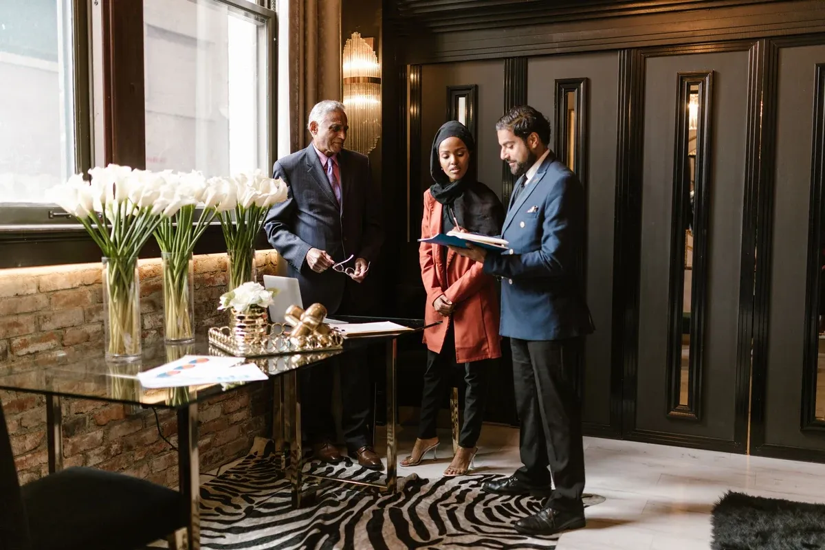 Three people in business attire review a folder beside a glass table with white calla lilies in a hall with dark paneled walls.