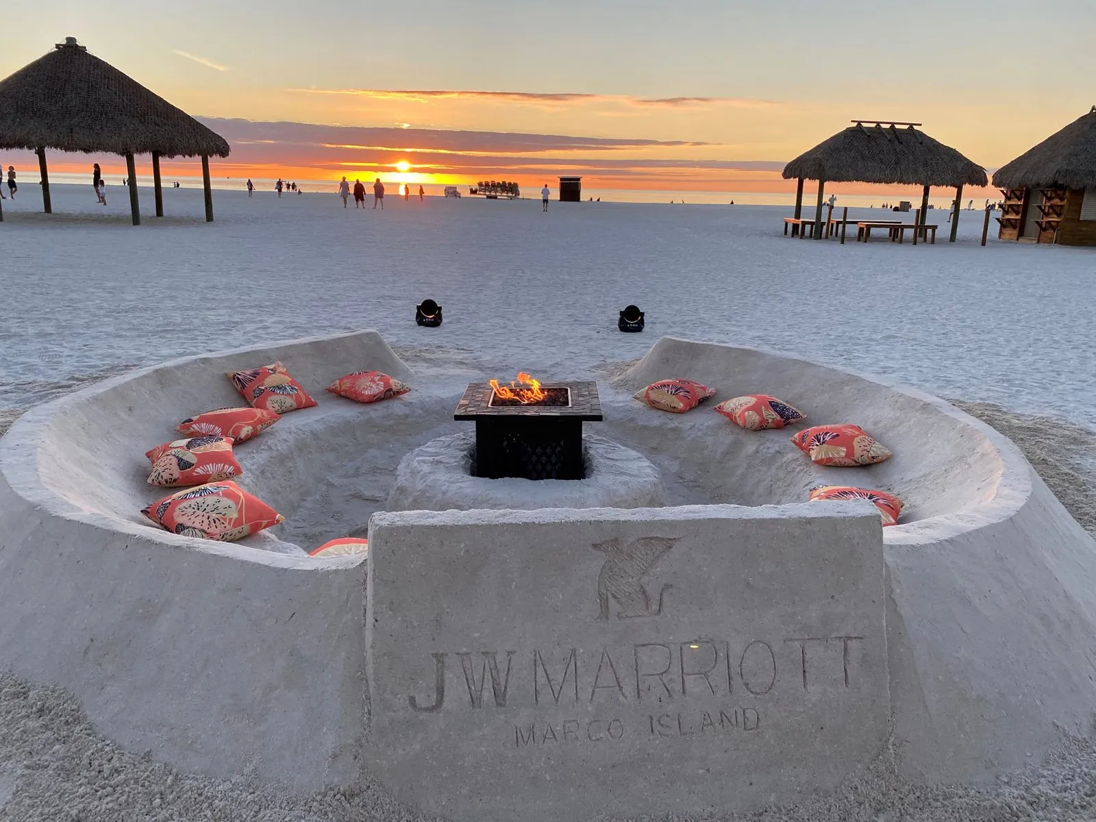 Sculpted sand lounge with cushions and a fire pit on the beach at sunset