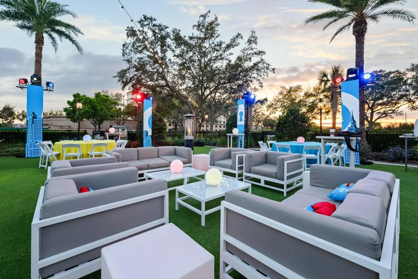 Outdoor lounge furniture on a lawn with palm trees and lit event signage at dusk