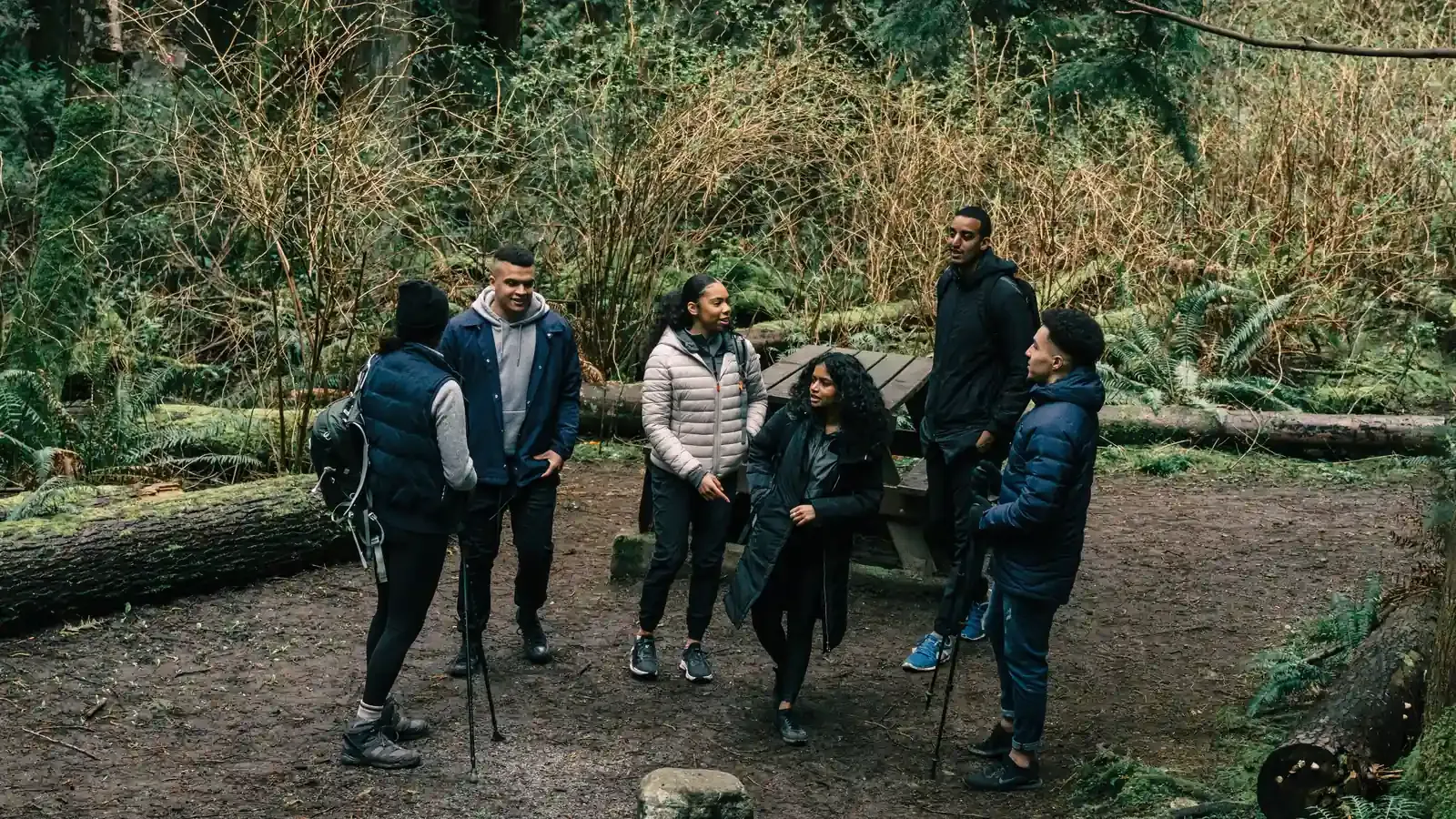 A group of six pauses on a forest trail beside a picnic table, chatting.