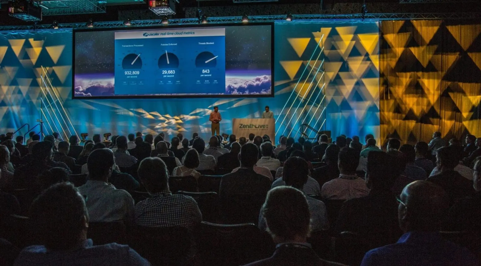 Conference keynote stage with a geometric backdrop and large screens