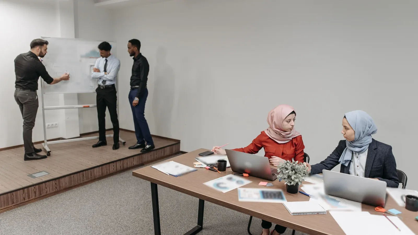 Three colleagues review a chart at a whiteboard on a low stage while two others work on laptops at a long table covered with printouts during a company offsite.