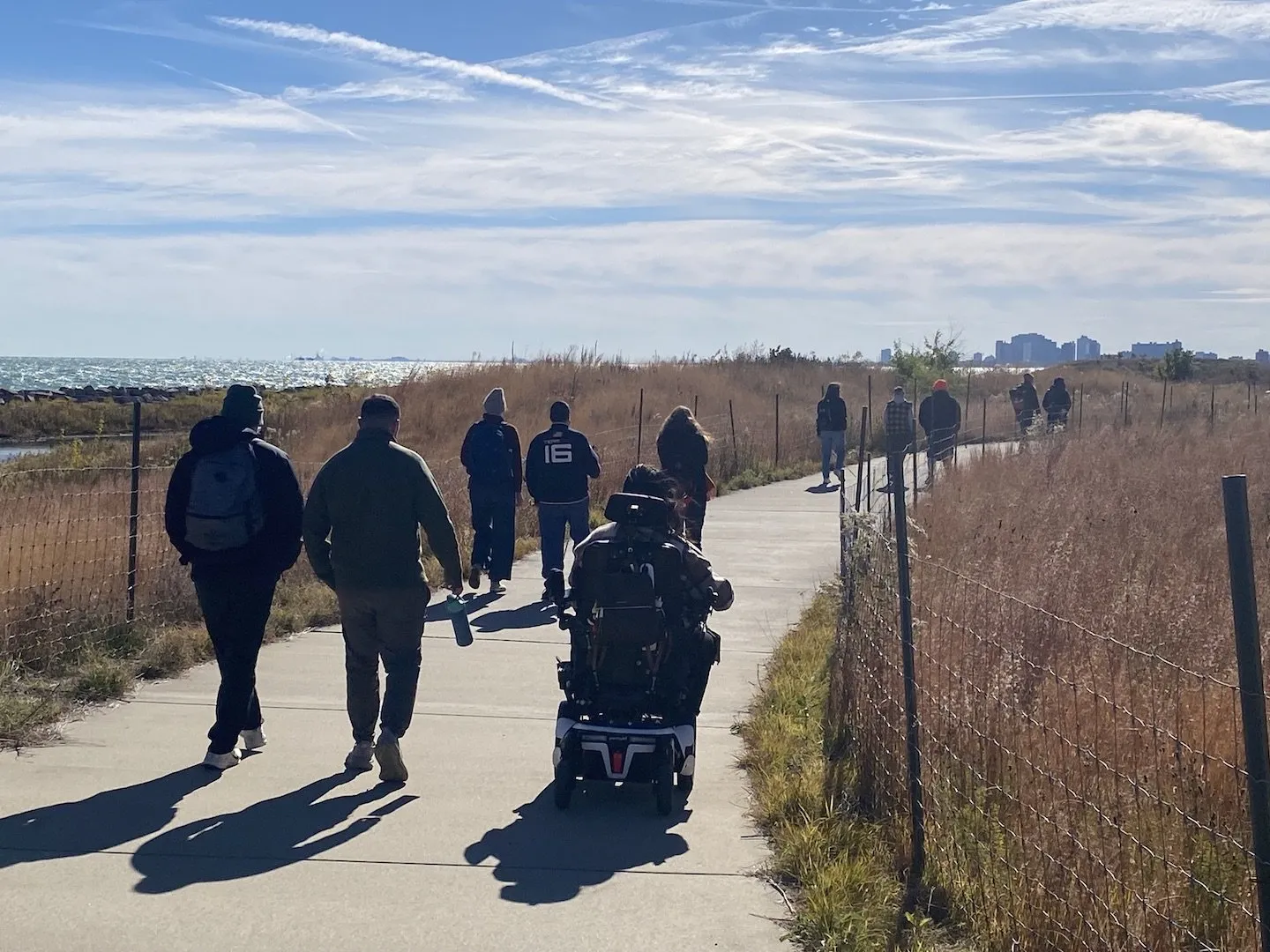 Group walking a lakefront prairie path with high-rises in the distance
